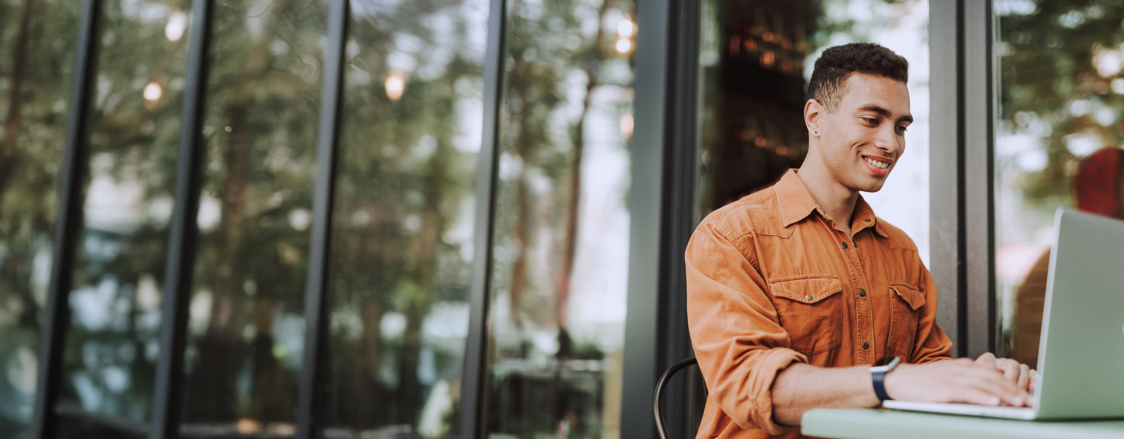 Junger Mann sitzt draußen im Café, arbeitet am Laptop und lächelt dabei.