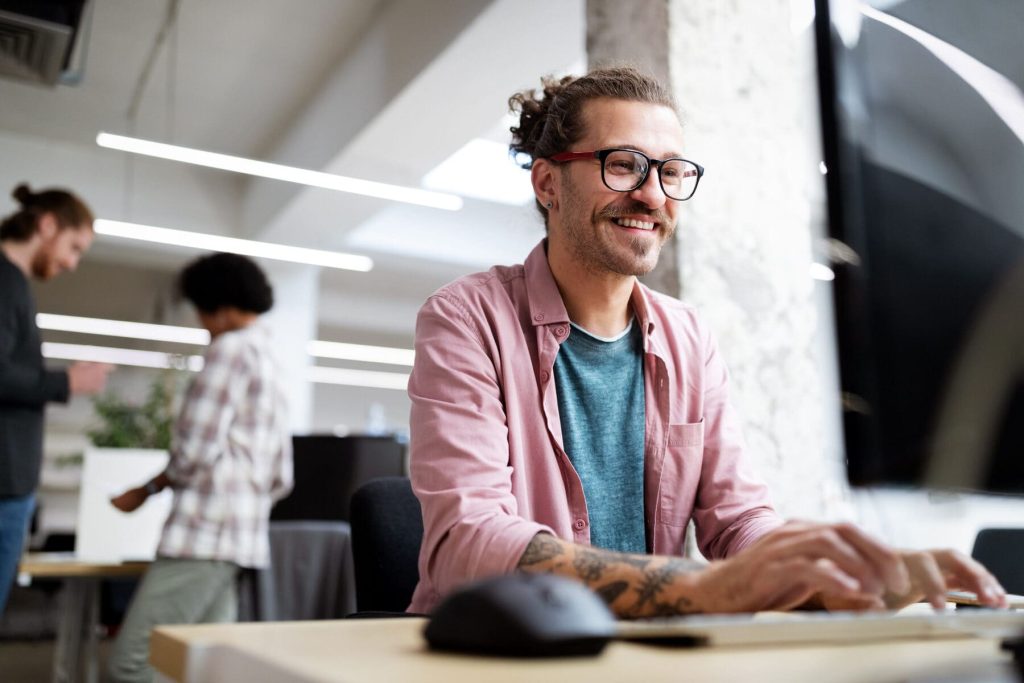 Junger Mann mit Brille und Tattoos arbeitet lächelnd am Computer in einem modernen Büro, im Hintergrund unterhalten sich zwei Kollegen